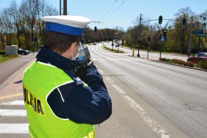 Umundurowany policjant stoi przy jezdni i trzymając w rękach urządzenie do pomiaru prędkości mierzy prędkość jadących ulicą samochodów.