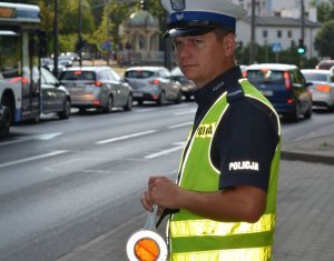 Umundurowany policjant stoi przy przy ulicy i obserwuje zachowania kierujących pojazdami.