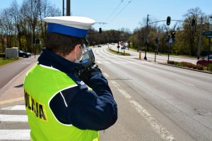 Umundurowany policjant ruchu drogowego trzyma w ręku urządzenie i mierzy prędkość jadących ulicą samochodów.