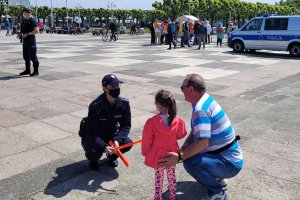 Umundurowana policjantka rozmawia na placu z dzieckiem i mężczyzną. Za nimi widoczny umundurowany policjant, radiowóz i inne osoby.