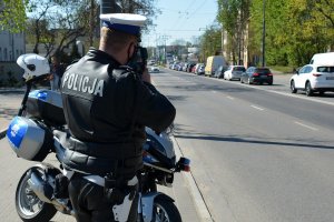 Umundurowany policjant mierzy prędkość pojazdów jadących ulicą. Za nim stoi oznakowany motocykl policyjny.