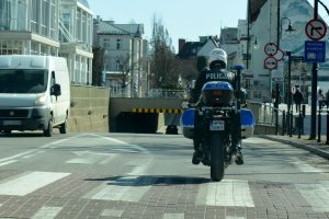 Umundurowany policjant jedzie ulicą na oznakowanym motocyklu. Pasem z naprzeciwka jedzie biały bus.