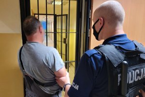 Umundurowany policjant prowadzi zatrzymanego mężczyznę. Za nimi widać kratę oraz korytarz znajdujący się w pomieszczeniu dla osób zatrzymanych.