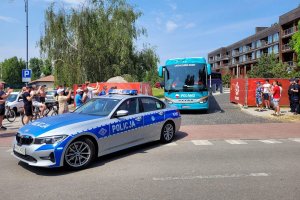 Oznakowany radiowóz eskortuje autokar, który za nim wyjeżdża z hotelu. Obok bramy stoją ludzie.