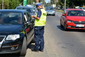 Umundurowana policjantka stoi przy ulicy i przeprowadza kontrolę drogową wobec kierującego pojazdem, który stoi na poboczu. Ulicą jadą samochody.