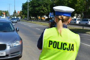 Umundurowana policjantka stoi przy jezdni i za pomocą urządzenia mierzy prędkość jadących ulicą pojazdów.