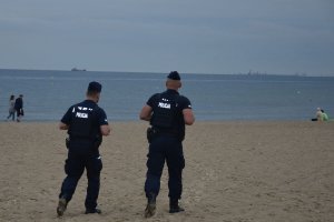 Umundurowani policjanci idą plażą. Za nimi w tle widać sylwetki osób, plaże i wodę.