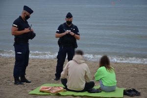 Plaża nad wodą. Umundurowani policjanci przeprowadzają interwencję wobec kobiety i mężczyzny siedzących na plaży.