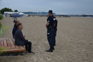 Plaża nad wodą. Umundurowani policjanci przeprowadzają interwencję wobec dwóch mężczyzn siedzących na plaży.