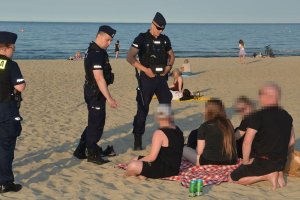 Umundurowani policjanci podejmują interwencję wobec czterech osób siedzących na kocu na plaży.