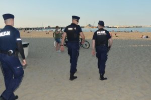 Trzech umundurowanych policjantów idzie po plaży. Za nimi widać osoby i wodę.