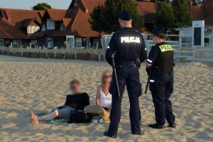 Dwóch umundurowanych policjantów rozmawia z osobami, które siedzą na kocu na plaży. Za nimi widać budynki od zabudowań przy plaży.