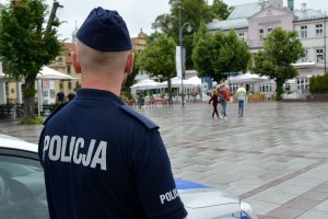 Umundurowany policjant stojąc przy radiowozie obserwuje osoby spacerujące po placu