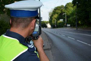 Fragment sylwetki umundurowanego policjanta, który za pomocą urządzenia mierzy prędkość jadących ulicą pojazdów.