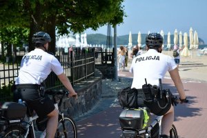 Dwóch umundurowanych policjantów na rowerach jedzie alejką nadmorską. Za nimi widać plażę, parasole, wodę i spacerujące osoby.