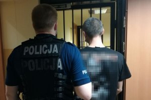 Korytarz w budynku. Umundurowany policjant prowadzi do pomieszczenia zatrzymanego mężczyznę.