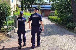 Umundurowani policjanci idą chodnikiem. Za nimi widoczny pociąg.