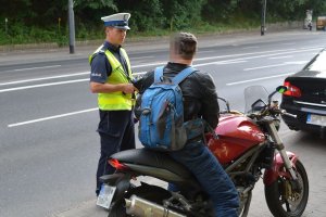 Umundurowany policjant kontroluje kierującego motocyklem. Obok nich stoi samochodów, a za nimi widoczna ulica i drzewa. - zdjęcie archiwalne