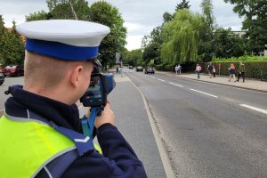 Umundurowany policjant stoi przy jezdni i przy pomocy urządzenia mierzy prędkość jadących pojazdów. W tle po chodniku idą piesi.