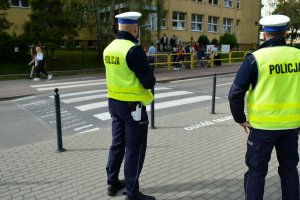 Umundurowani policjanci stoją przy przejściu dla pieszych. Za nimi po drugiej stronie ulicy widoczni ludzie i budynek szkoły.