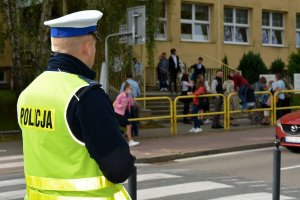 Umundurowany policjant stoi przy przejściu dla pieszych. Za nim po drugiej stronie ulicy widoczni ludzie i budynek szkoły.