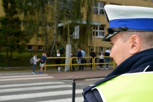 Umundurowany policjant stoi przy przejściu dla pieszych. Za nim po drugiej stronie ulicy widoczni ludzie i budynek szkoły.