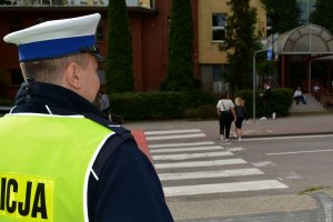 Umundurowany policjant stoi przy przejściu dla pieszych, po którym idą osoby.