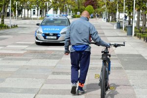 Mężczyzna prowadzi rower po deptaku. Za nim z naprzeciwka jedzie oznakowany radiowóz z włączonymi sygnałami świetlnymi