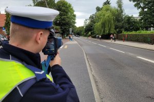 Umundurowany policjant stoi przy jezdni i za pomocą urządzenia mierzy prędkość pojazdów poruszających się po drodze. W oddali widać osoby idące po chodnikach.