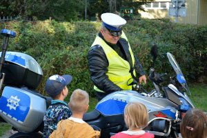 Teren posesji. Umundurowany policjant stoi przy oznakowanym motocyklu. Przed nim tyłem stoi czworo dzieci.