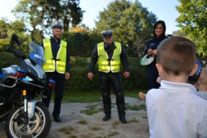 Ogrodzony teren. Policjanci Umundurowani stoją przy radiowozie. Przed nimi stoi dziecko i patrzy w ich stronę.