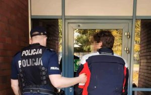 Umundurowany policjant prowadzi zatrzymanego mężczyznę do budynku.