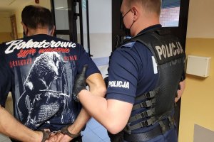 Policjant prowadzi w stronę drzwi zatrzymanego mężczyznę, który ma założone na ręce trzymane z tyłu kajdanki