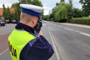 Umundurowany policjant stoi przy ulicy i za pomocą urządzenia trzymanego w rękach mierzy prędkość jadących samochodów.