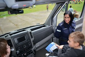 Umundurowana policjantka rozmawia z dziećmi, które siedzą na fotelach kierowcy  i pasażera radiowozu. Za nimi widać grupy innych dzieci