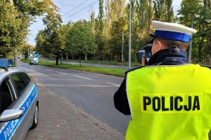 Umundurowany policjant stoi przy ulicy, po której jadą samochody i za pomocą urządzenia trzymanego w rękach mierzy ich prędkość. Obok policjanta widać na poboczu fragment oznakowanego radiowozu.