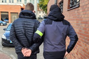 Chodnik przed budynkiem. Policjant w stroju cywilnym i z założoną na lewym ramieniu żółtą opaską z napisem POLICJA prowadzi zatrzymanego mężczyznę, który ma założone kajdanki na ręce trzymane z tyłu.