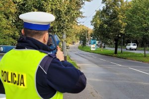 Umundurowany policjant stoi przy jezdni i za pomocą urządzenia trzymanego w rękach mierzy prędkość jadących ulicą pojazdów