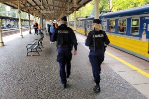 Umundurowani policjanci idą wzdłuż peronu dworca. Po prawej stronie na peronie stoi pociąg, a w tle widać osoby.