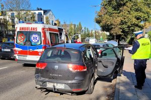 Umundurowany policjant stoi przy zatoczce autobusowej i wykonuje czynności na miejscu zderzenia dwóch pojazdów. W tle widoczny ambulans, ulica i jadący po niej samochód.