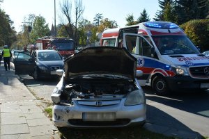 Umundurowany policjant i strażacy wykonują czynności na miejscu zderzenia dwóch pojazdów. Obok na jezdni widoczny ambulans.