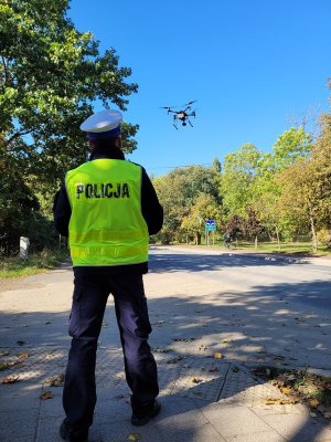 Umundurowany policjant stoi przy jezdni. Przed nim w powietrzu widoczny dron.