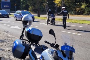 Umundurowany policjant stoi na jezdni i zatrzymuje do kontroli kierującego motocyklem. Za nimi widoczne inne pojazdy. Przy jezdni stoi oznakowany motocykl policyjny.