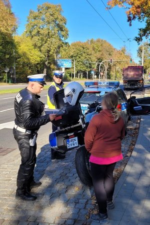 Umundurowani policjanci rozmawiają z kobietą. Obok nich oznakowany motocykl i radiowóz. Za nimi w zatoczce stoi pojazd ciężarowy, a obok ulica.