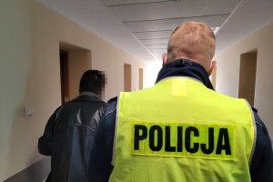 Korytarz. Umundurowany policjant prowadzi zatrzymanego mężczyznę.