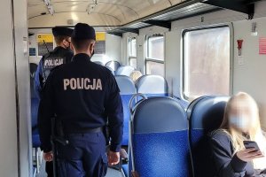 Dwaj umundurowani policjanci idą przez przedział pociągu. Na siedzeniach widoczne sylwetki osób.