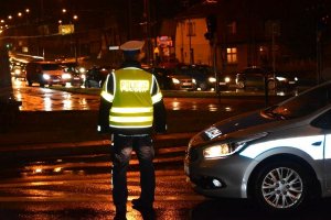 Pora nocna. Umundurowany policjant stoi przy ulicy i obserwuje jadące ulicą pojazdy. Obok policjanta widoczny fragment oznakowanego radiowozu.