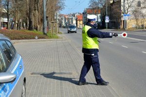 Umundurowany policjant stoi na jezdni. W jednej ręce trzyma tarczę do zatrzymywania pojazdów, a drugą ręką pokazuje kierującemu pojazdem, że to o niego chodzi. Za policjantem widoczny samochód, który jedzie w stronę policjanta.