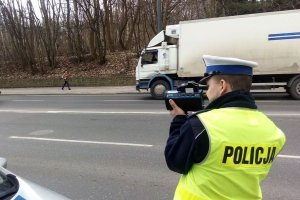 Umundurowany policjant stoi przy jezdni i trzyma w rękach urządzenie do pomiaru prędkości. Za nim widoczny pojazd ciężarowy, który jedzie ulicą i las.