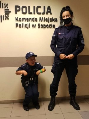 Umundurowana policjantka stoi z małym chłopcem ubranym w strój policyjny przy ścianie, na której widnieje napis KOMENDA MIEJSKA POLICJI W SOPOCIE